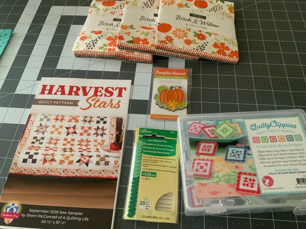 An assortment of quilting supplies including the 'Harvest Stars' quilt pattern booklet, packs of 'Birch & Willow' fabric, a 'Pumpkin Harvest' enamel needle minder, Clover flower-shaped pins, and a box of 'QuiltyClippies' fabric clips.