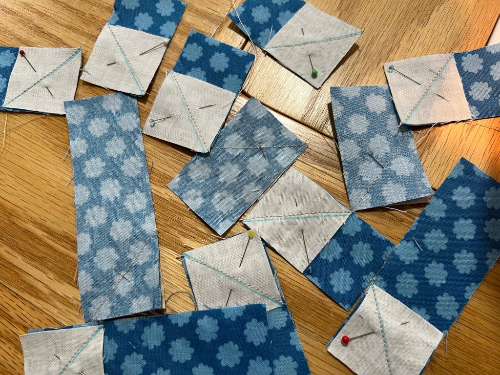 A collection of fabric pieces in various shades of blue, some with floral patterns, arranged on a wooden surface. The pieces are pinned together, with visible stitching and threads.