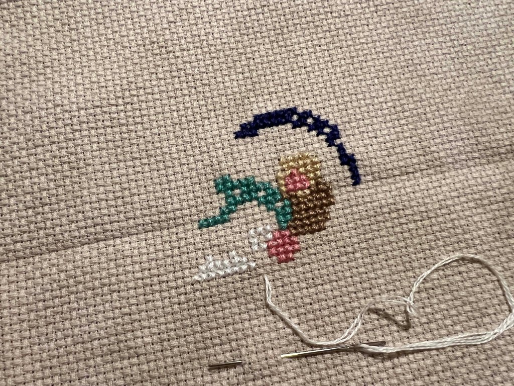 Close-up of an unfinished embroidery piece on beige fabric, featuring colorful stitched flowers and a crescent shape, with a needle and thread visible.